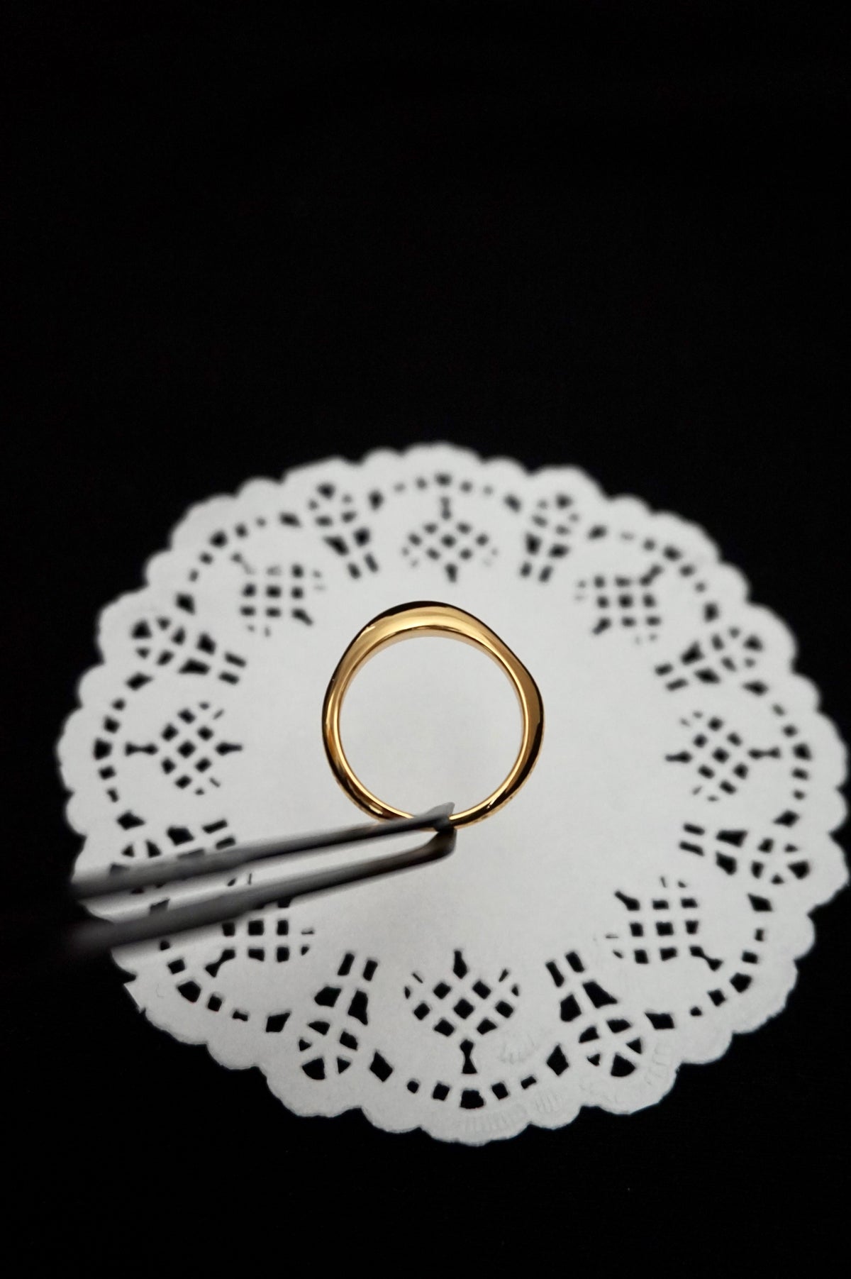 minimal everyday gold ring on a white doily with tweezers against a black background