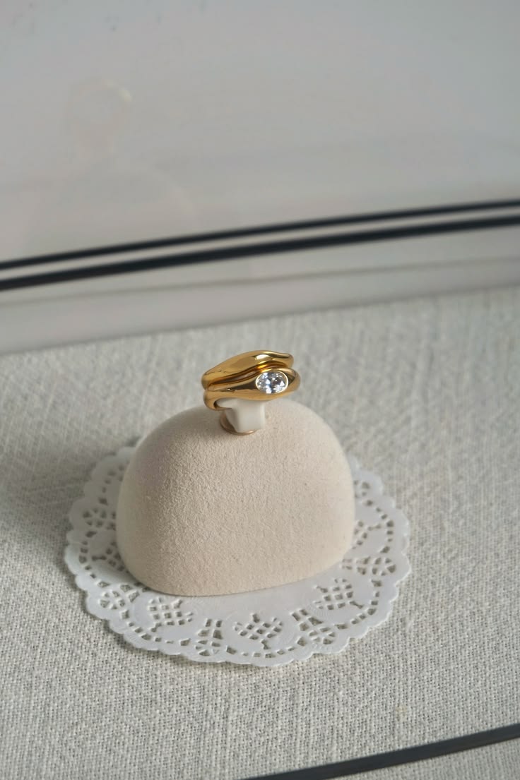 Gold ring with a diamond on a small white cushion with lace edge, placed on a textured surface.