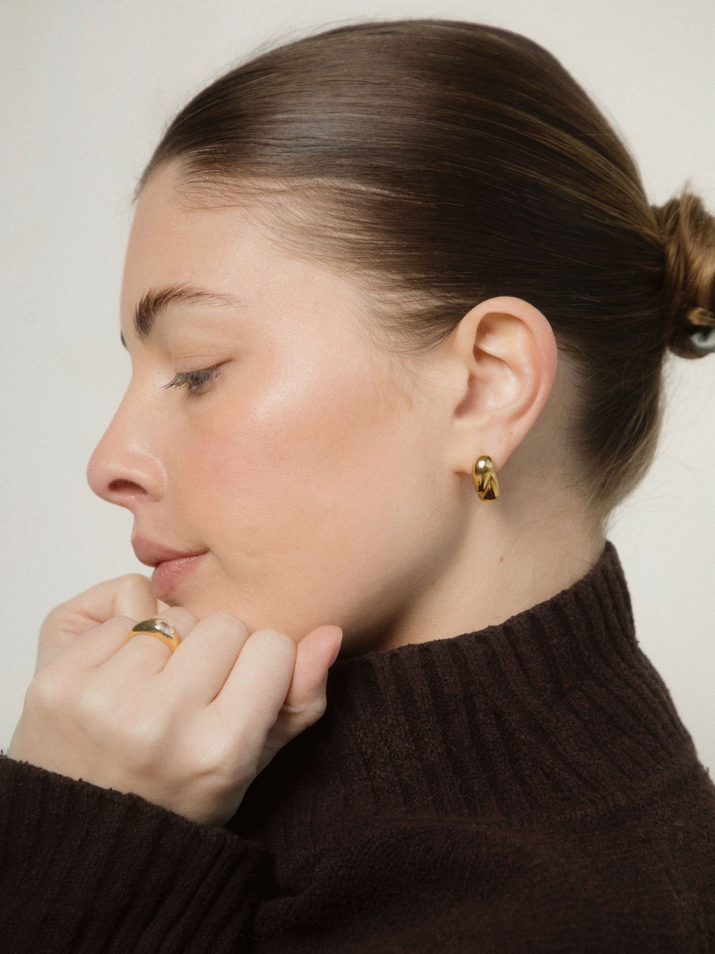Woman wearing gold earrings and a ring, with a neutral background