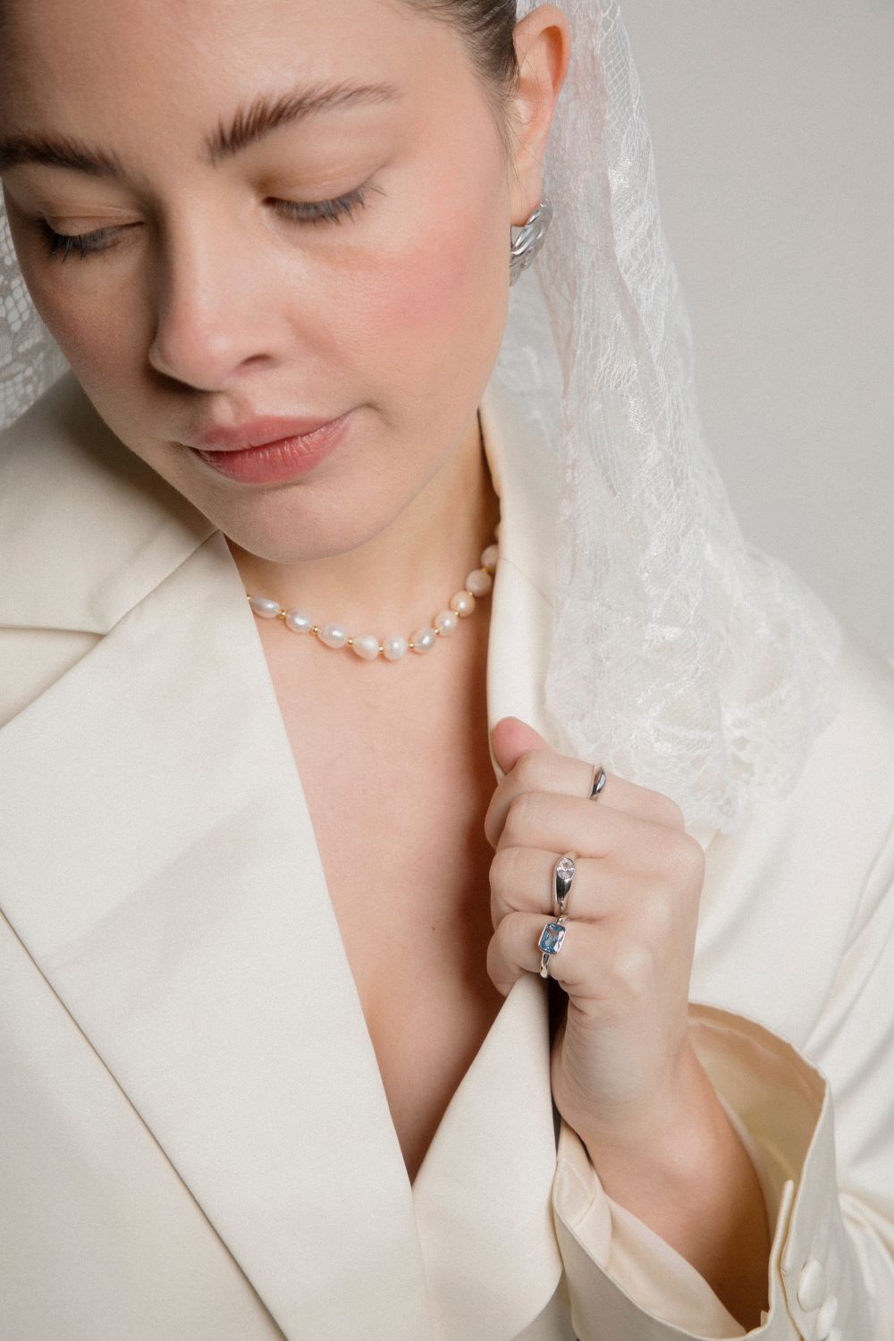 Women wearing pearl necklace in a beige blazer with lace headscarf. Showcasing on her fingers are the Skye Ring and Something Blue Ring from Third Tone jewelry brand