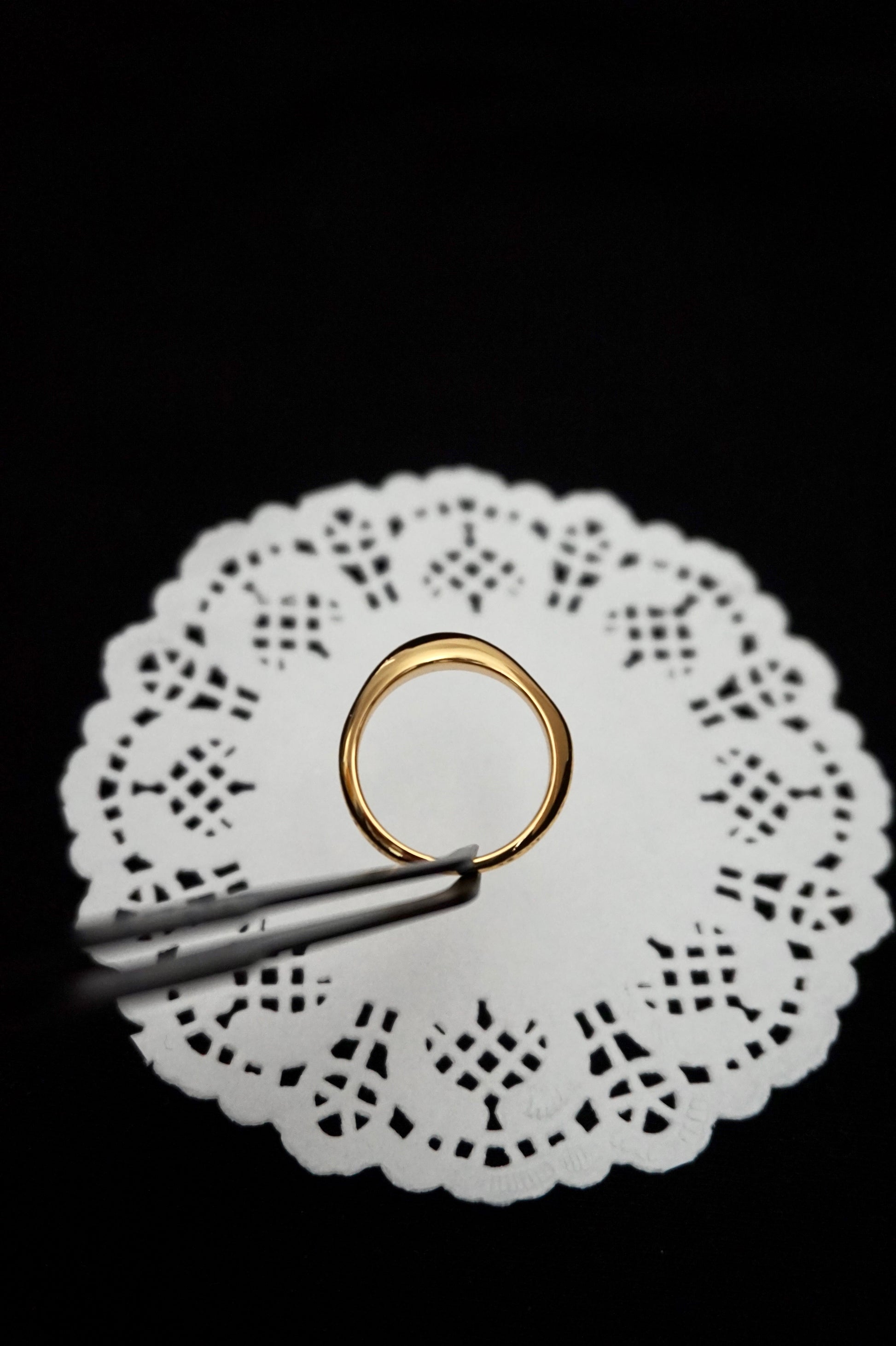 minimal everyday gold ring on a white doily with tweezers against a black background